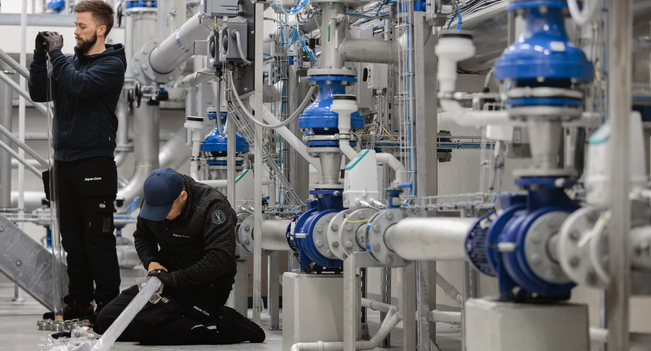 Två servicetekniker från Aqua & Care arbetar med installation och inspektion av UV-komponenter i ett avancerat vattenreningssystem omgivet av rör och ventiler.